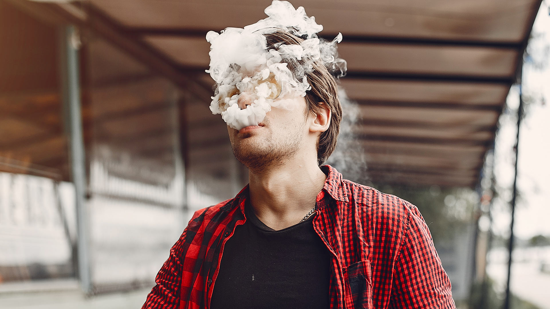 A young man in a red and black plaid shirt vaping with thick smoke obscuring his face, amidst an ambiguous airport background, relevant to the blog topic 'Can You Vape at the Airport